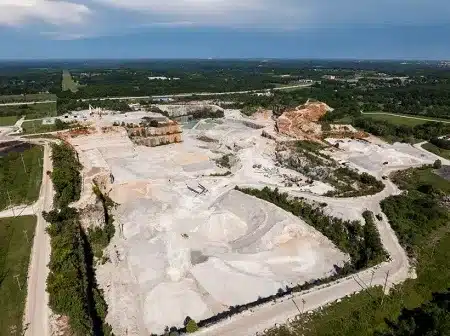 Aerial view of an open-pit quarry with exposed white limestone and terraced benches, surrounded by roads and forests.