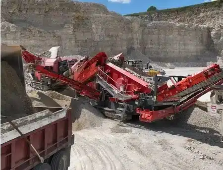 Red rock-crushing conveyor system in a quarry, with a dump truck in the foreground and tall rocky cliffs in the background.