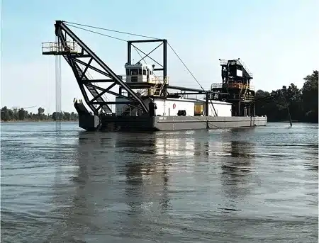 Large dredging barge with towering crane on a calm river/harbor, blue sky overhead